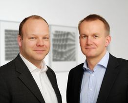 Zwei Männer in Anzügen stehen nebeneinander, im Hintergrund sind unscharfe Bilder an der Wand.