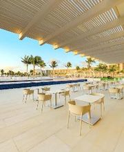 Terraza al aire libre con mesas y sillas bajo pérgola, vista de piscina y palmeras al fondo.