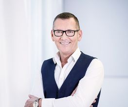 Ein lächelnder Mann mit Brille und Anzugweste steht vor einem hellen Hintergrund.
