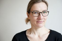 Frau mit Brille und schwarzem Hemd lächelt freundlich in die Kamera vor grauem Hintergrund.