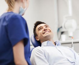Patient smiling at dentist, sitting in a dental chair, with dentist in blue scrubs and a mask.