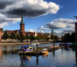 Flussansicht mit Booten, Stadtgebäuden und Kirchen unter wolkigem Himmel.