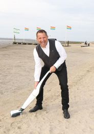 Ein Mann in Anzug saugt Sand am Strand, im Hintergrund wehen bunte Fahnen im Wind.