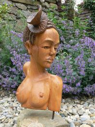 Holzskulptur einer Frau vor lila Blumen und Steinen im Garten.