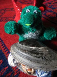 Grünes Plüschdrache mit Muschel und Wollknäuel auf rotem Teppich.