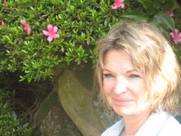 Eine Frau lächelt in der Sonne, umgeben von grünen Büschen und rosa Blüten.