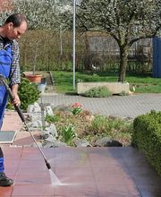 Mann reinigt Terrasse mit Hochdruckreiniger im Garten, trägt blaue Latzhose und kariertes Hemd.