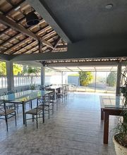 Área externa coberta, com mesas e cadeiras de ferro, vegetação ao fundo e placa de Wi-Fi na parede.