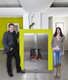 Zwei Personen stehen neben einem modernen, silbernen Kamin vor einer leuchtend grünen Wand.