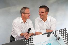 Zwei Männer in weißen Hemden lächeln und lehnen auf einem Schild mit Mikrofonmotiv.