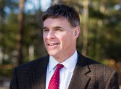 Man in a suit and red tie standing outdoors, trees blurred in the background.