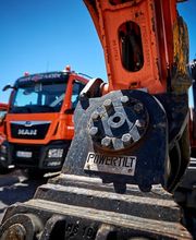 Orange Baggerarm mit "Powertilt"-Schriftzug, im Hintergrund drei rote Lastwagen auf Baustelle.