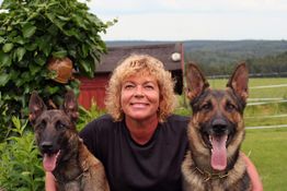 Eine Person mit lockigem Haar posiert mit zwei großen Hunden im Freien vor einer grünen Landschaft.