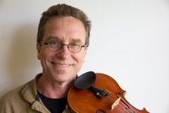 Person lächelt, hält Geige an der Schulter, trägt Brille, freundlicher Ausdruck, heller Hintergrund.