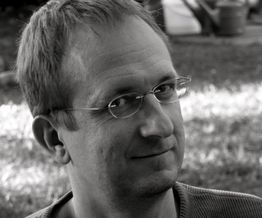 Mann mit Brille lächelt in die Kamera, schwarz-weiß Foto, im Freien aufgenommen.