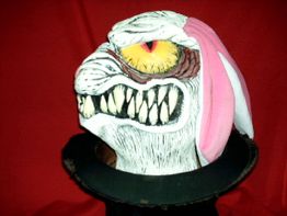 Gruselige Maske mit gelben Augen, scharfen Zähnen und pinken Hasenohren vor rotem Hintergrund.