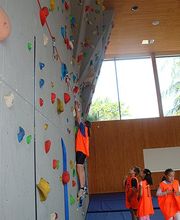 Kinder klettern in einer Turnhalle eine bunte Kletterwand hoch.