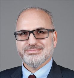 Man with glasses and a beard wearing a suit and blue shirt against a gray background.