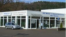 Autowerkstatt mit Glasfront und silbernem Auto davor auf einem Parkplatz.