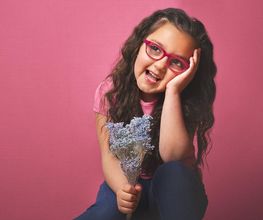 Niña con gafas rosadas sonríe y sostiene flores en fondo rosa.
