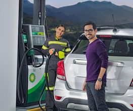 Hombre sonriente junto a su coche en una gasolinera con una empleada, montañas de fondo.