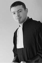 Un homme en robe d'avocat noire et blanche, regarde la caméra, fond gris neutre.