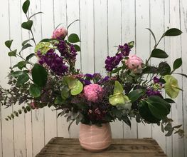 Ramo de flores variadas en jarrón rosado sobre mesa de madera, fondo de pared blanca de madera.