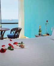 Habitación con cama blanca, mesa con sillas, rosas y vista al mar a través de una gran ventana.