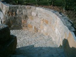 Steintreppe führt in eine runde Vertiefung mit gemauerter Steinwand, umgeben von Grünpflanzen.