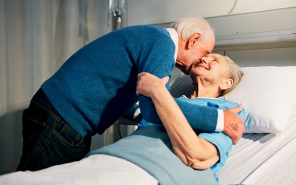 Älterer Mann umarmt und küsst eine ältere Frau, die in einem Krankenhausbett liegt.