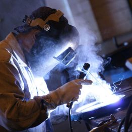 Welder working with bright sparks, wearing protective gear and mask in a dimly lit workshop.