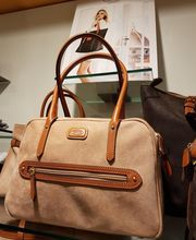 Beige Handtasche mit braunen Griffen, ausgestellt auf einem Regal vor einem Poster.