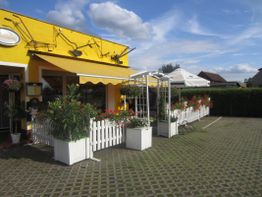 Gelbes Gebäude mit Terrasse, weißen Zäunen und Blumenarrangements unter blauem Himmel.