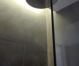 Ducha con cabezal circular y azulejos oscuros, iluminada cálidamente desde arriba.