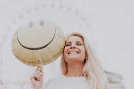 Smiling woman holding a yellow smiley balloon with a ferris wheel in the blurry background.