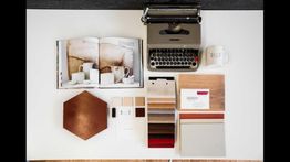 Macchina da scrivere, libro aperto, tazza e campioni di materiali su un tavolo bianco.